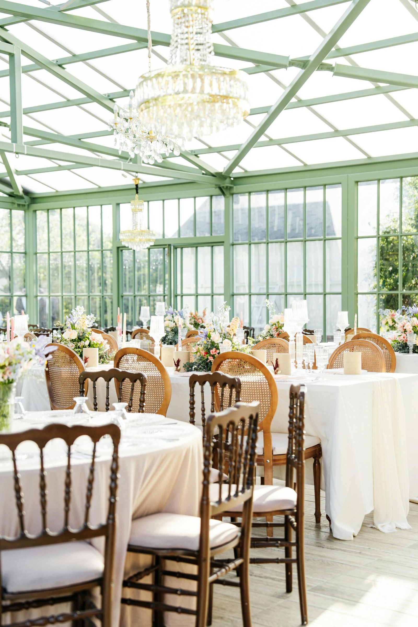 Sunlit glass venue set for an elegant wedding reception with floral decorations and chandeliers.